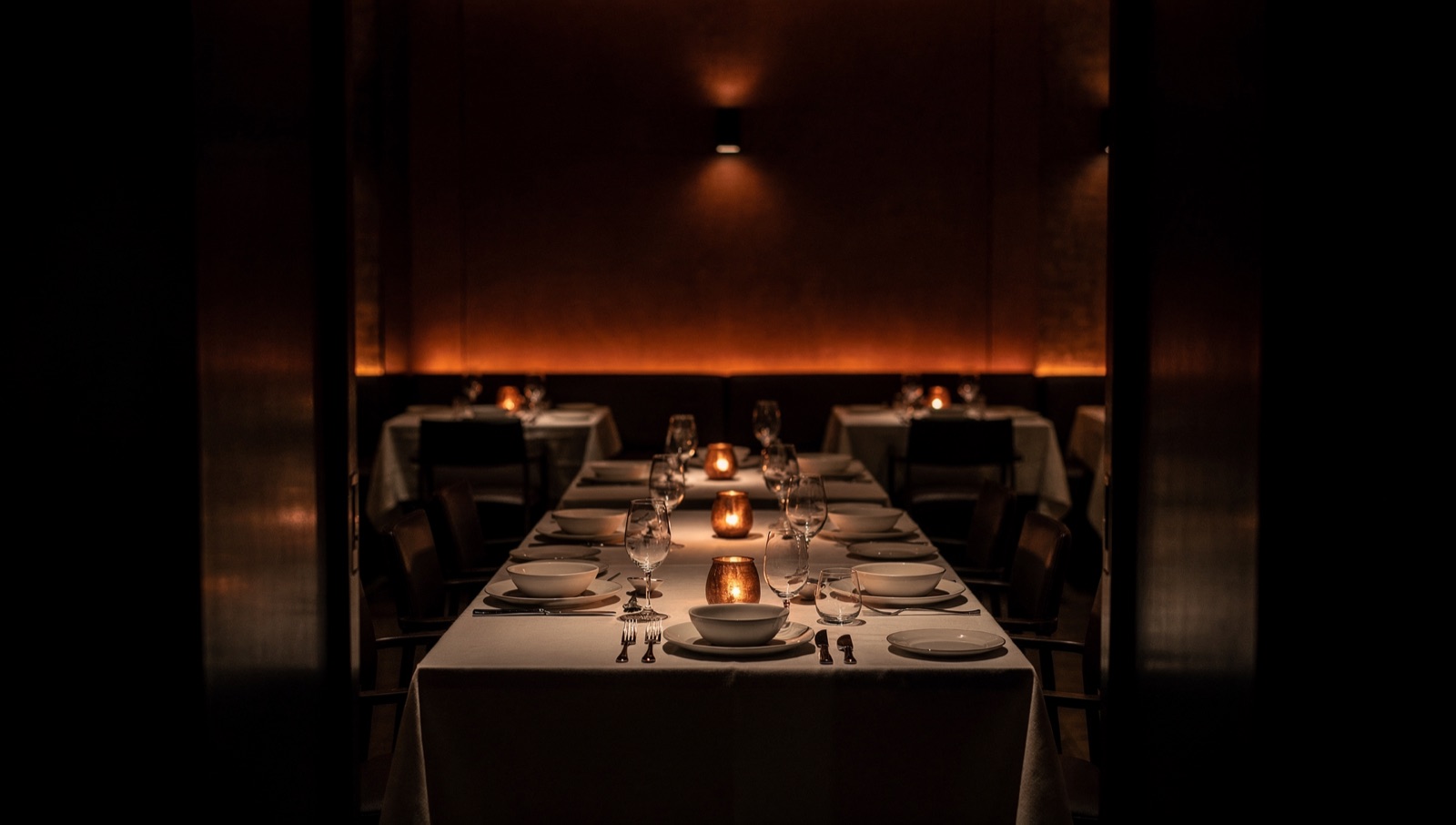Candlelit dining room receding into warm darkness, copper light on linen tables.
